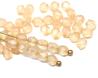 May include: Peach-colored faceted beads, strung on gold-colored wire, are scattered across a white background. The beads have a subtle, translucent quality, and are arranged in rows. A few loose beads are also visible.