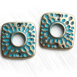 Puede incluir: Dos colgantes cuadrados de metal con acabado bronce envejecido y detalles turquesa. Cada colgante presenta una abertura circular central y un patrón de pequeños agujeros irregulares en los bordes. Para la creación de joyas o manualidades.