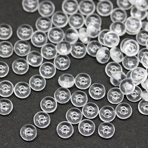 Puede incluir: Una colección de pequeños cierres de presión de plástico transparente esparcidos sobre una superficie oscura. Cada cierre tiene un diseño circular con un agujero central y un mecanismo de presión. Los cierres son translúcidos.