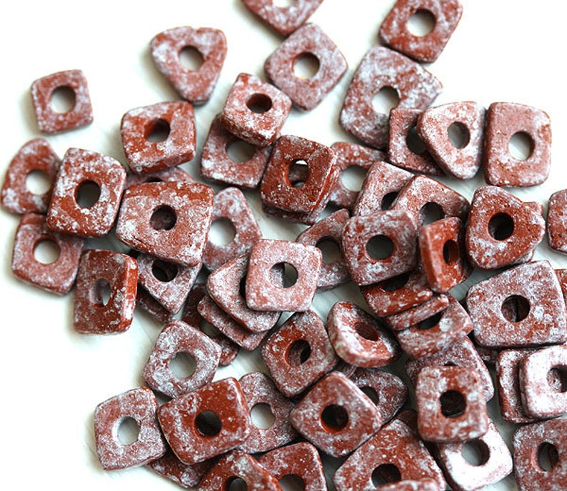 May include: A pile of red and white ceramic square beads with a hole in the centre. The beads have a speckled, textured surface.