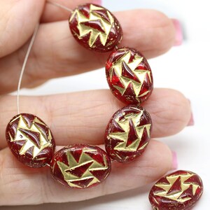 May include: A strand of oval-shaped glass beads with a red base and gold geometric pattern. The beads are strung on a thin white cord.