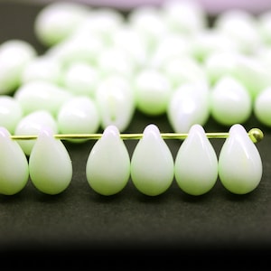 May include: Close-up of pale green teardrop-shaped beads. The beads are strung on a thin gold wire, with several more beads out of focus in the background. The beads have a pearlescent finish, suggesting they are made of glass or a similar material.