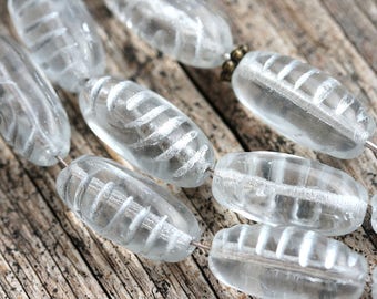 May include: A strand of clear glass beads with a white swirl design. The beads are oval shaped and have a smooth, polished finish.