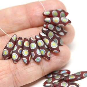 May include: A strand of maroon glass beads with white and iridescent circles. The beads are shaped like teardrops and are strung on a silver wire.