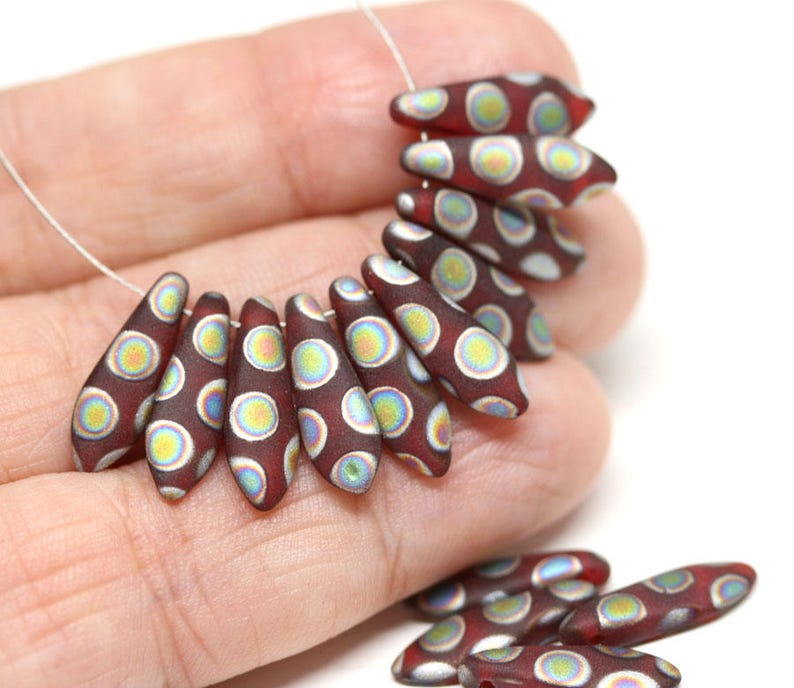 May include: A strand of maroon glass beads with white and iridescent circles. The beads are shaped like teardrops and are strung on a silver wire.