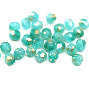 May include: A collection of faceted, turquoise-colored beads with gold flecks. These beads are scattered across a white surface, showcasing their irregular shapes and translucent quality. Ideal for jewelry making or craft projects.