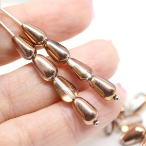 Può includere: Una fila di perline di vetro color rame a forma di lacrima. Le perline hanno un bagliore metallico e sono infilate su un filo d'argento.