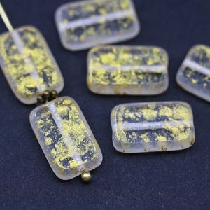 Pode incluir: Contas retangulares de vidro transparente com inclusões de folha de ouro. As contas têm um padrão dourado salpicado. Algumas são enfiadas com pequenas contas castanhas escuras e um alfinete dourado. Em um fundo azul escuro.