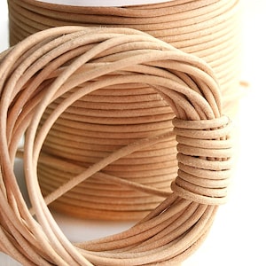 Pode incluir: Close-up de dois rolos de cordão de couro castanho claro. Um rolo está no fundo e o outro em primeiro plano, com uma parte do cordão desenrolada. O cordão é enrolado firmemente e a cor é um bege natural.