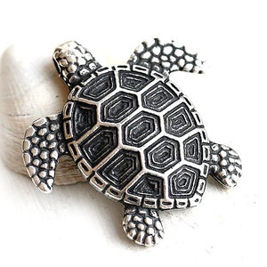 May include: A silver turtle pendant with a detailed shell design. The turtle is facing right and has a textured surface.