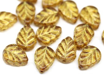 Puede incluir: Una colección de cuentas de vidrio en forma de hoja de color ámbar. Cada cuenta presenta un diseño detallado de venas de hoja en un tono dorado más oscuro. Las cuentas están dispersas sobre una superficie blanca, mostrando sus detalles.
