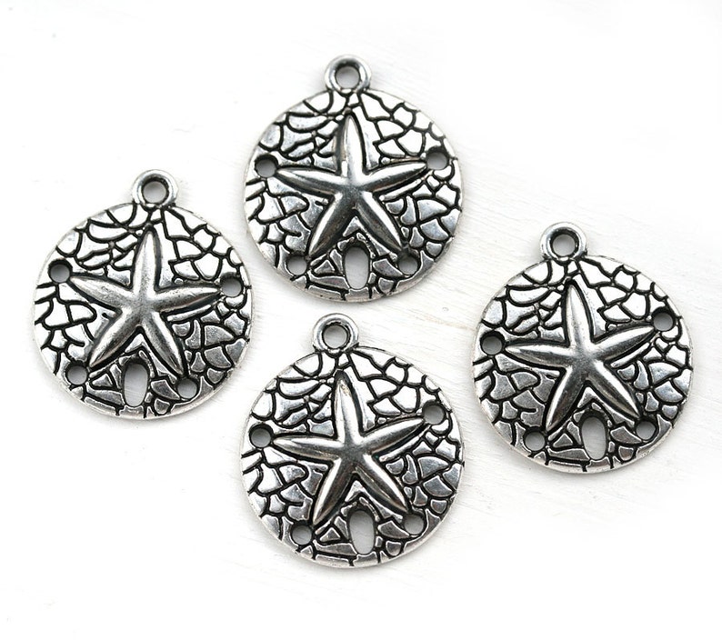 May include: Four silver-toned metal charms shaped like sand dollars with a starfish design in the center. Each charm has three holes around the edge.