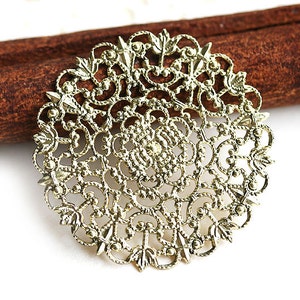 May include: Gold filigree metal charm with a circular design. The charm has a delicate, intricate pattern with a floral motif.