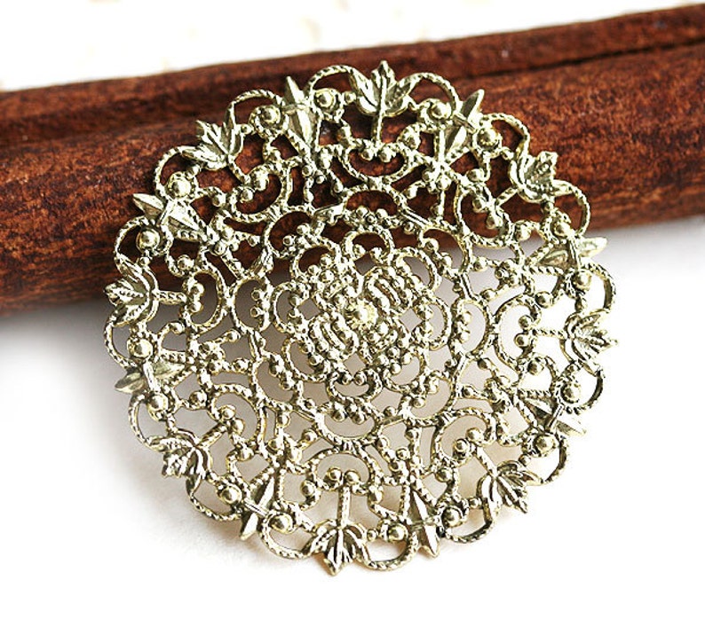 May include: Gold filigree metal charm with a circular design. The charm has a delicate, intricate pattern with a floral motif.