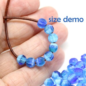 Peut inclure: Un fil de perles de verre bleues et blanches sur un cordon marron. Les perles sont rondes et stri&eacute;es. Le texte "size demo" est visible. Un tas de perles similaires est dans le coin inf&eacute;rieur droit.