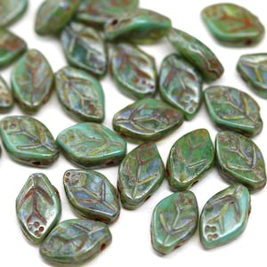 May include: A collection of leaf-shaped beads in shades of green, with intricate vein and berry details. The beads have a glossy finish and are scattered across a white surface, ideal for jewelry making or craft projects.