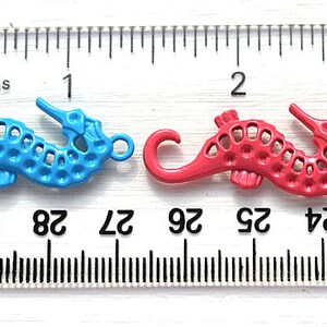 May include: Two metal seahorse charms, one blue and one red, are shown on a ruler. The ruler is marked in inches and sixteenths of an inch. The seahorses are about 1 inch long.