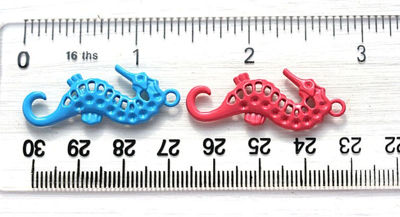 May include: Two metal seahorse charms, one blue and one red, are shown on a ruler. The ruler is marked in inches and sixteenths of an inch. The seahorses are about 1 inch long.