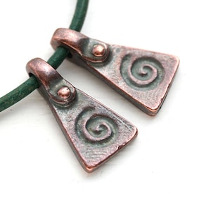 May include: Two copper pendants with a spiral design hang from a green leather cord. The pendants are triangular in shape and have a textured surface.