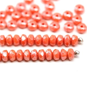 May include: Close-up of coral-colored beads. The beads are faceted and have a glossy finish. Some beads are strung together, while others are loose. The beads are a vibrant orange-pink color and are set against a white background.