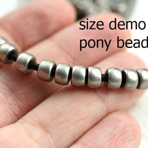 May include: Close-up of a silver pony bead necklace on a black cord. The beads are small and round, and the cord is thin. The text "size demo pony beads" is visible in the image.
