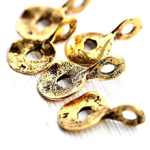 May include: Close-up of several antique gold-colored teardrop-shaped jewelry charms. Each charm has a textured surface, a circular hole, and a loop at the top. The charms are scattered on a white surface.