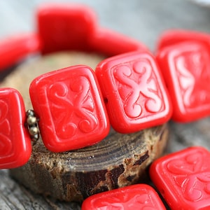 Könnte beinhalten: Nahaufnahme eines roten Armbands. Die quadratischen Perlen weisen ein geschnitztes Blumenmuster auf. Das Armband ist mit goldfarbenem Metall verbunden. Die Perlen sind etwa 2 cm groß.