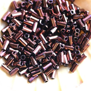 Könnte beinhalten: Ein Haufen kleiner, dunkelbrauner Glasperlen mit einem metallischen Schimmer. Die Perlen sind zylindrisch geformt und haben eine leicht irisierende Oberfläche.