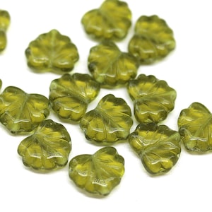 Peut inclure: Un ensemble de perles en verre vert olive en forme de feuilles. Chaque perle présente un motif de veines de feuilles détaillé. Les perles sont dispersées sur une surface blanche, créant une esthétique naturelle. Idéal pour la création de bijoux et l'artisanat.