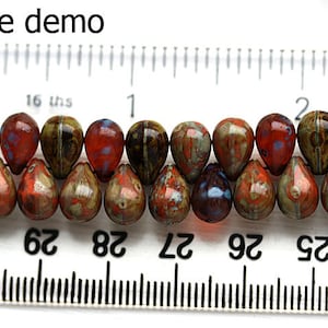 Puede incluir: Primer plano de una cadena de 14 cuentas de vidrio en forma de l&aacute;grima. Las cuentas son una mezcla de marr&oacute;n, naranja y rojo con un efecto moteado, marmoleado. Las cuentas est&aacute;n enhebradas en un alambre y se miden con una regla. La regla muestra que las cuentas tienen aproximadamente 2,5 cm de largo.