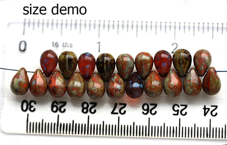 Puede incluir: Primer plano de una cadena de 14 cuentas de vidrio en forma de l&aacute;grima. Las cuentas son una mezcla de marr&oacute;n, naranja y rojo con un efecto moteado, marmoleado. Las cuentas est&aacute;n enhebradas en un alambre y se miden con una regla. La regla muestra que las cuentas tienen aproximadamente 2,5 cm de largo.