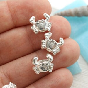Puede incluir: Tres abalorios de plata en forma de cangrejo, cada uno con un dise&ntilde;o detallado, se muestran en una mano. Los abalorios son peque&ntilde;os y delicados, perfectos para a&ntilde;adir un toque de fantas&iacute;a a las joyas o las manualidades.