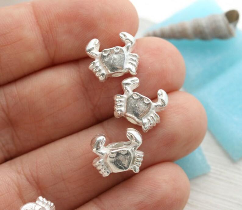 Puede incluir: Tres abalorios de plata en forma de cangrejo, cada uno con un dise&ntilde;o detallado, se muestran en una mano. Los abalorios son peque&ntilde;os y delicados, perfectos para a&ntilde;adir un toque de fantas&iacute;a a las joyas o las manualidades.