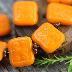 Op de afbeelding: Een streng oranje glazen kralen met een bloemmotief. De kralen zijn vierkant en hebben een glanzende afwerking.