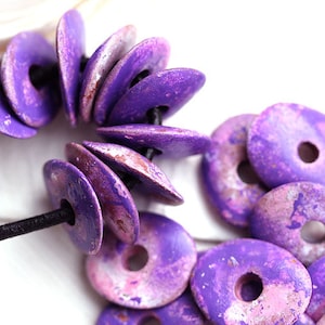 Può includere: Primo piano di perline di legno viola con finitura anticata. Le perline sono a forma di disco con un foro al centro, infilate su un cordino nero. Il colore è un viola vibrante con sfumature di rosa e marrone.