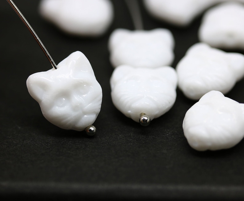 May include: White ceramic cat face beads with a hole for stringing. The beads are smooth and have a glossy finish.
