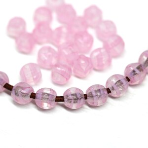 Peut inclure: Des perles de verre roses avec un motif ray&eacute;, enfil&eacute;es sur un cordon marron. Les perles sont rondes et translucides, avec une lumi&egrave;re douce et diffuse. Quelques perles en vrac sont dispers&eacute;es en arri&egrave;re-plan.