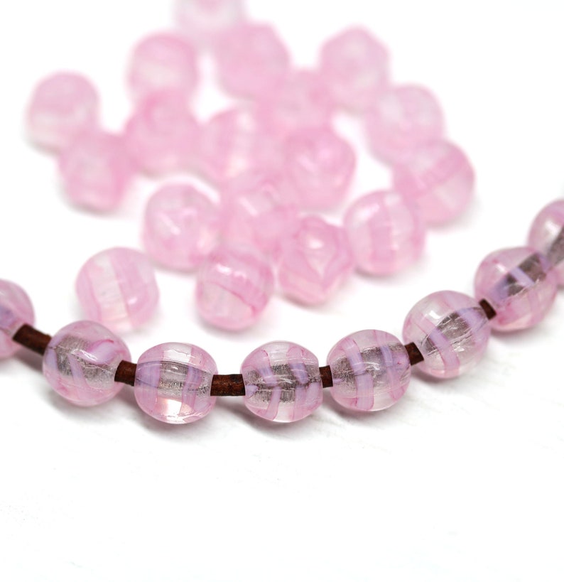 Peut inclure: Des perles de verre roses avec un motif ray&eacute;, enfil&eacute;es sur un cordon marron. Les perles sont rondes et translucides, avec une lumi&egrave;re douce et diffuse. Quelques perles en vrac sont dispers&eacute;es en arri&egrave;re-plan.