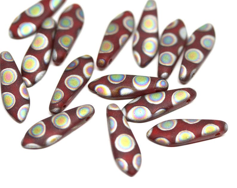 May include: A group of 15 red glass beads with iridescent silver and green accents. The beads are shaped like long, narrow ovals and have a pattern of white circles on them.
