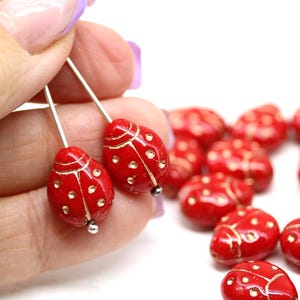 Peut inclure: Boucles d'oreilles coccinelles rouges avec des détails dorés et des crochets argentés. Les boucles d'oreilles ont une finition brillante. D'autres perles coccinelles sont visibles en arrière-plan.