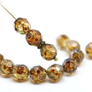 Peut inclure: Gros plan sur des perles ambrées à facettes avec un motif moucheté. Les perles sont enfilées sur un fil doré, avec quelques perles nettes et d'autres floues en arrière-plan. Les perles ont un aspect texturé unique.