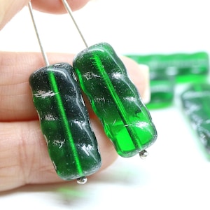 May include: Two rectangular, translucent emerald green glass beads with a wavy, textured surface and a vertical hole through the center. Each bead features a distinct, deep green hue and a slightly irregular, organic shape with subtle silver-toned accents.