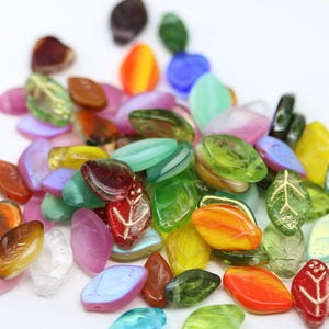 Può includere: Una collezione di perline a forma di foglia di vetro colorate in varie tonalità di verde, giallo, arancione, rosso, viola e blu. Alcune perline presentano un disegno a venature di foglie. Le perline sono sparse su una superficie bianca.