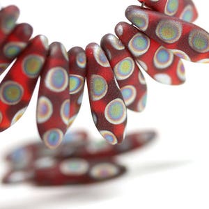 May include: A close-up of red and white glass beads with a speckled pattern. The beads are shaped like teardrops and have a iridescent finish.