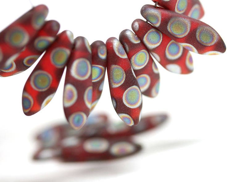 May include: A close-up of red and white glass beads with a speckled pattern. The beads are shaped like teardrops and have a iridescent finish.