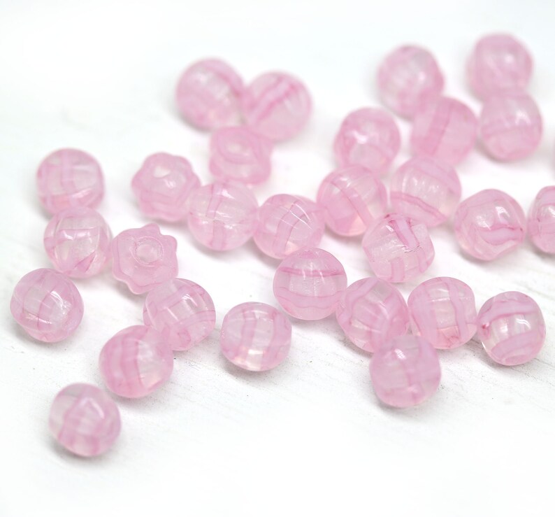 Peut inclure: Un ensemble de perles roses translucides avec un motif ray&eacute;. Les perles sont rondes, certaines ayant une forme florale. Elles sont dispos&eacute;es sur une surface blanche, cr&eacute;ant une esth&eacute;tique d&eacute;licate et f&eacute;minine. Id&eacute;al pour la cr&eacute;ation de bijoux.