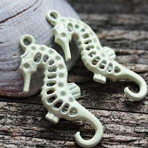 May include: Two light green metal seahorse charms with a cutout design. The seahorses are hanging from a hook.