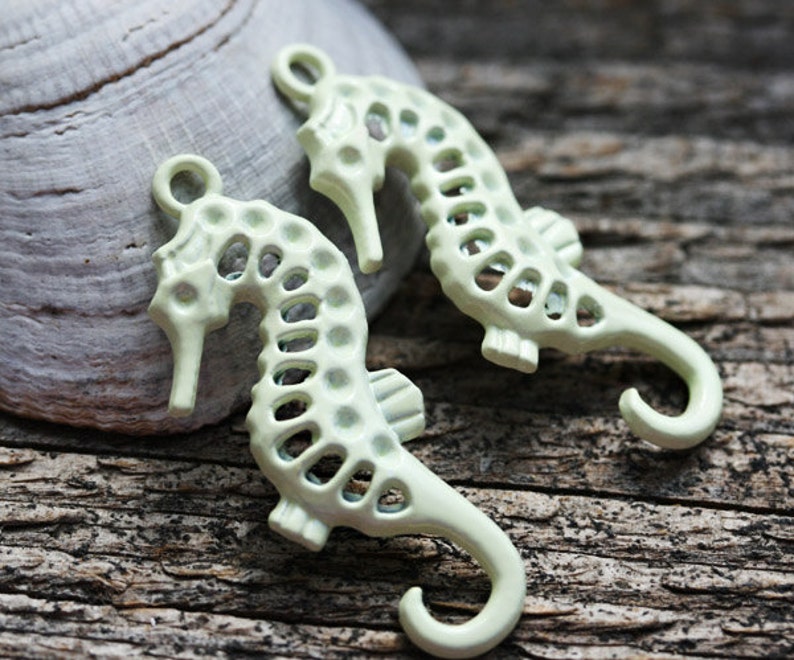 May include: Two light green metal seahorse charms with a cutout design. The seahorses are hanging from a hook.