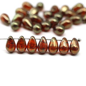 Op de afbeelding: Druppelvormige kralen in koper-, brons- en olijfgroene tinten. De kralen zijn geregen op een dunne draad, waarbij sommige scherp en andere onscherp zijn. De kralen hebben een metallic glans. Elke kraal is ongeveer 1 cm lang.