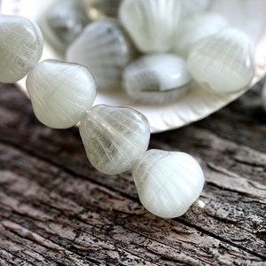 Può includere: Una collana di perline di vetro bianco a forma di conchiglie. Le perline hanno una superficie testurizzata e sono infilate su un filo d'argento.